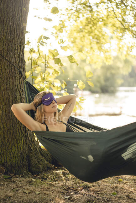 Young woman relaxing in hammock by the lake, wearing sleeping mask — Stock Photo