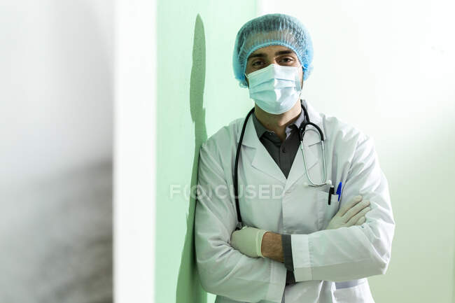 Medico maschio con cappuccio chirurgico in piedi con le braccia incrociate in ospedale durante COVID-19 — Foto stock