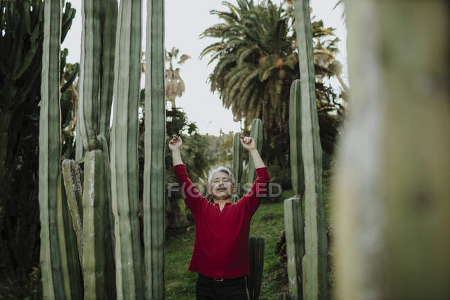 Uomo maturo con le braccia alzate in piedi tra le piante di cactus in giardino — Foto stock