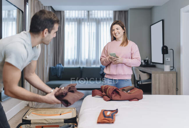 Smiling woman looking at man unpacking luggage in hotel room — Stock Photo