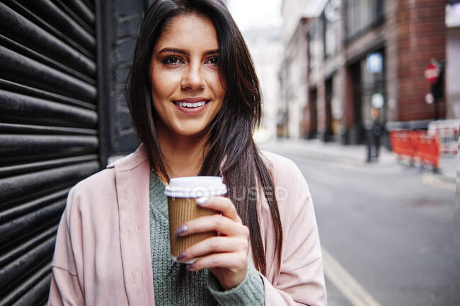 Усміхнена молода жінка з одноразовою чашкою кави стоїть за затвор у місті — стокове фото
