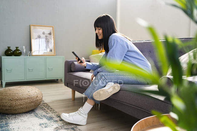 Mujer sonriente usando un teléfono inteligente mientras está sentada en el sofá en la sala de estar en casa - foto de stock