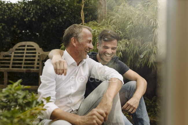 Cheerful son with arm around father sitting in backyard — Stock Photo