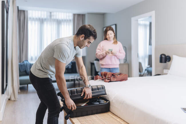 Hombre adulto medio desempacando el equipaje en la cama con la mujer en segundo plano en el hotel — Stock Photo
