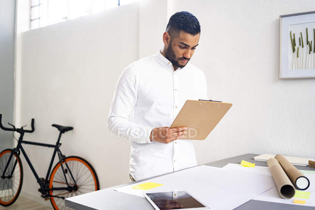 Чоловік-підприємець тримає буфер обміну, стоячи біля столу біля стіни в офісі — стокове фото