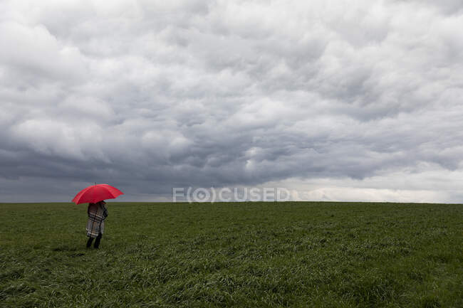 Femme avec parapluie rouge debout dans l'herbe par temps orageux — Photo de stock