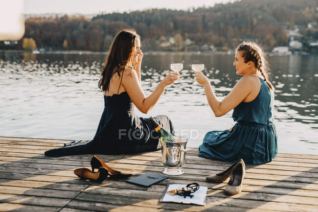 Female event planners toasting champagne while sitting on jetty over lake — Stock Photo