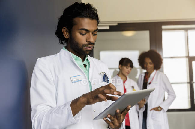 Junge männliche Gesundheitsfachangestellte mit digitalem Tablet, während weibliche Kollegen im Hintergrund im Krankenhaus stehen — Stockfoto