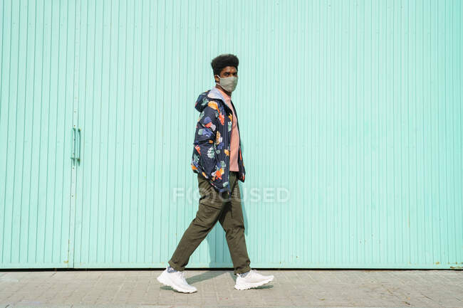 Homme avec les mains dans les poches portant un masque protecteur tout en marchant par mur turquoise — Photo de stock