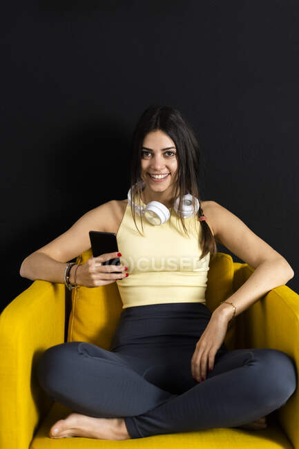 Femme sportive souriante avec écouteurs et téléphone portable assis sur un fauteuil à la maison — Photo de stock