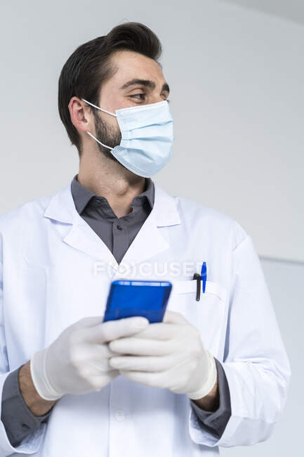 Мужчина медицинский эксперт носит защитную маску для лица, держа смартфон, отводя взгляд — стоковое фото