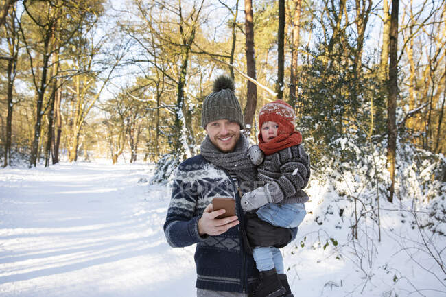 Padre con teléfono inteligente llevando hijo en la nieve durante el invierno - foto de stock