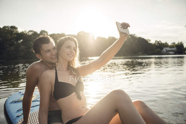 Счастливая пара, сидящая на доске на озере, фотографируя смартфоны — стоковое фото