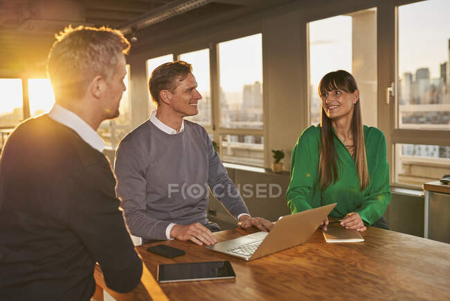 Homme d'affaires avec ordinateur portable souriant tout en étant assis par des collègues au bureau — Photo de stock