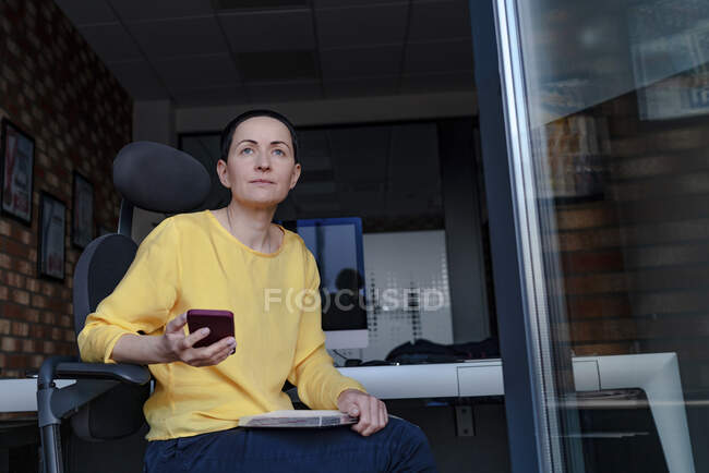 Thoughtful female entrepreneur with mobile phone looking away while on chair in modern offic — Stock Photo
