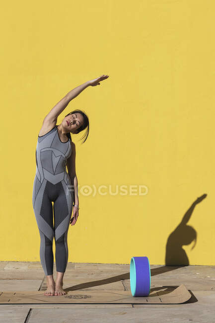 Mujer con los brazos levantados haciendo ejercicio con rueda de yoga contra pared amarilla - foto de stock