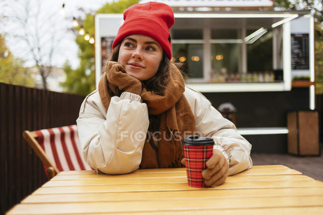 Frau in warmer Kleidung schaut weg, während sie mit der Hand am Kinn im Straßencafé sitzt — Stockfoto