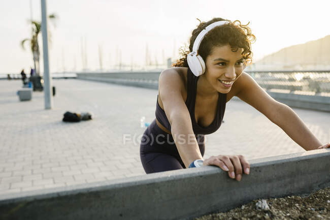Sportiva sorridente che ascolta musica attraverso le cuffie mentre si esercita sul ponte — Foto stock