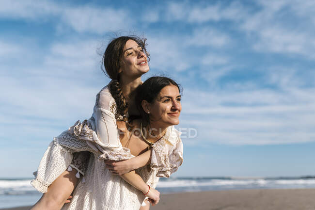 Young woman looking away while piggybacking female friend at beach — Stock Photo