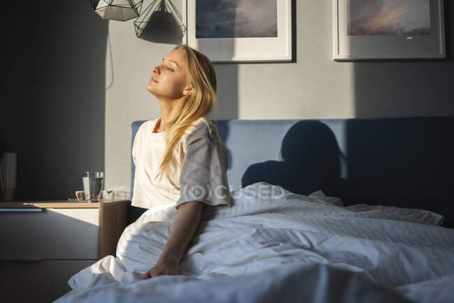 Lumière du soleil sur la femme assise avec les yeux fermés sur le lit à la maison — Photo de stock