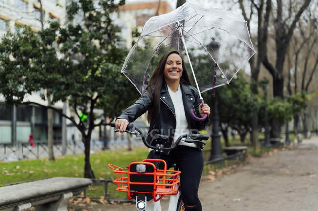 Belle femme en vélo électrique tout en tenant un parapluie dans la rue en ville — Photo de stock