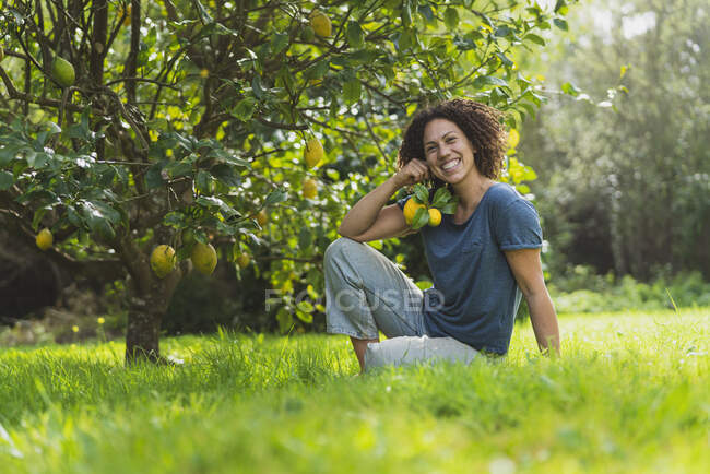 Donna felice che tiene limoni mentre siede sull'erba sotto l'albero di limone — Foto stock