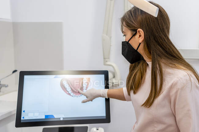 Dentista feminina usando máscara protetora apontando para a tela do computador na clínica — Fotografia de Stock