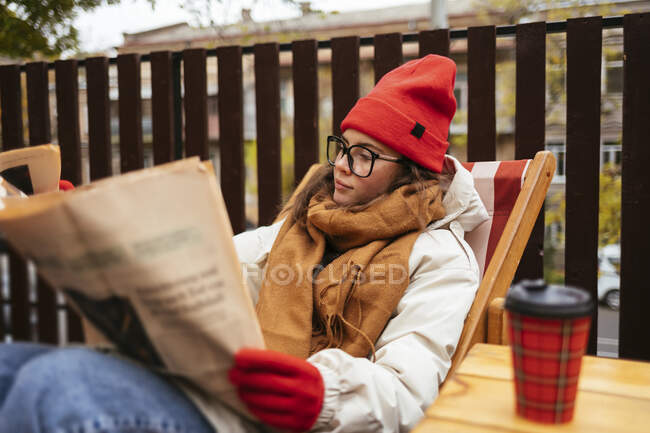 Jeune femme en vêtements chauds lecture journal tout en se relaxant sur la chaise au café — Photo de stock