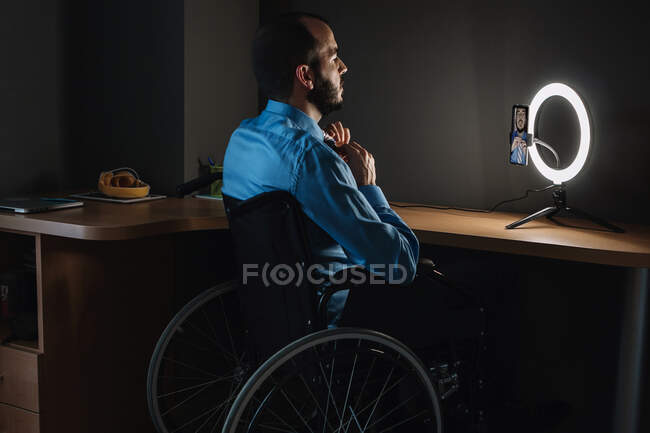 Инвалид-блоггер вяжет галстук во время видеосвязи через мобильный телефон, сидя на инвалидной коляске в студии — стоковое фото