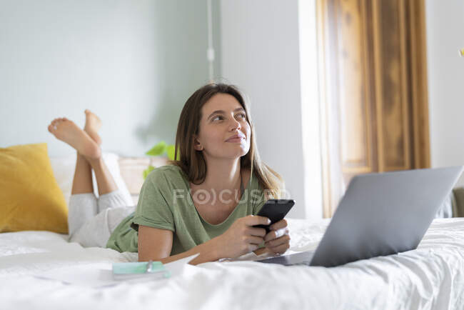 Вважна молода жінка з розумним телефоном лежить перед ноутбуком на ліжку. — стокове фото