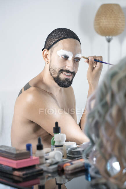 Uomo sorridente che applica ombretto bianco davanti allo specchio a casa — Foto stock
