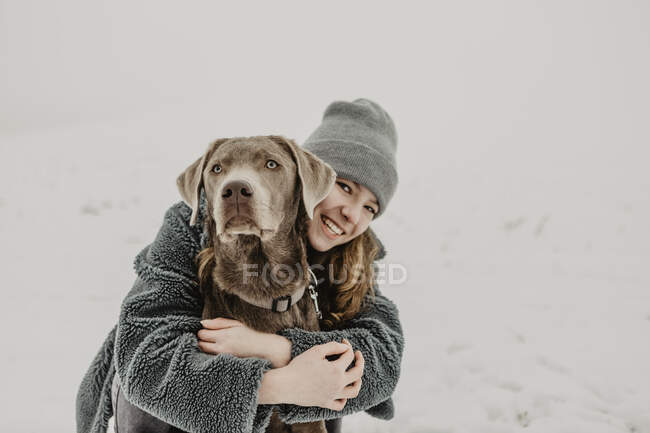 Porträt eines Teenagers, der im Schnee hockt und Labrador Retriever umarmt — Stockfoto