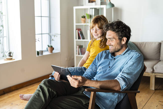 Padre e hija mirando la tableta digital mientras están sentados en el sillón en casa - foto de stock