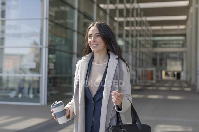 Femme entrepreneure souriante tenant sac à main et tasse jetable tout en détournant les yeux — Photo de stock