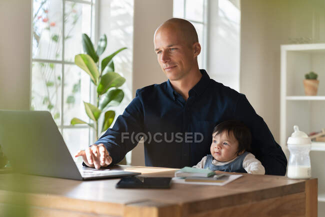 Мужчина-предприниматель работает на ноутбуке, держа ребенка в домашнем офисе — стоковое фото