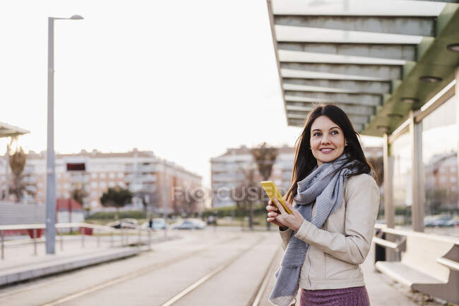 Souriant belle femme tenant téléphone intelligent tout en regardant loin de la gare — Photo de stock