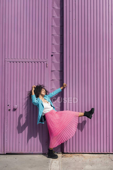 Игривая молодая женщина танцует у фиолетовой стены хижины в солнечный день — стоковое фото