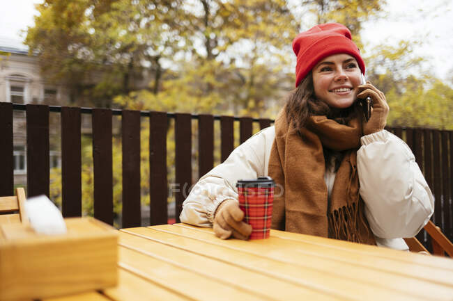 Jovem sorrindo enquanto falava no celular sentado no café da calçada — Fotografia de Stock