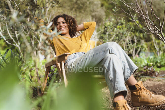 Mujer sonriente mirando hacia otro lado mientras se relaja en la silla durante la primavera en el huerto - foto de stock