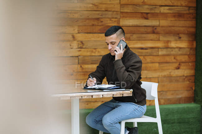 Young male entrepreneur talking on the phone while writing in diary at sidewalk cafe — Stock Photo