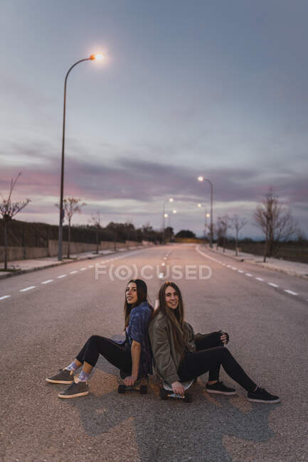 Amis souriantes assis dos à dos sur le skateboard pendant le crépuscule — Photo de stock
