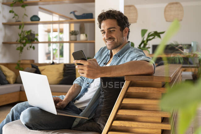 Lavoratore freelance maschio sorridente con computer portatile utilizzando smart phone mentre seduto divano in soggiorno — Foto stock