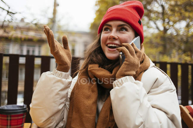 Mujer sonriente con sombrero de punto haciendo gestos mientras está sentada en la acera cafetería - foto de stock