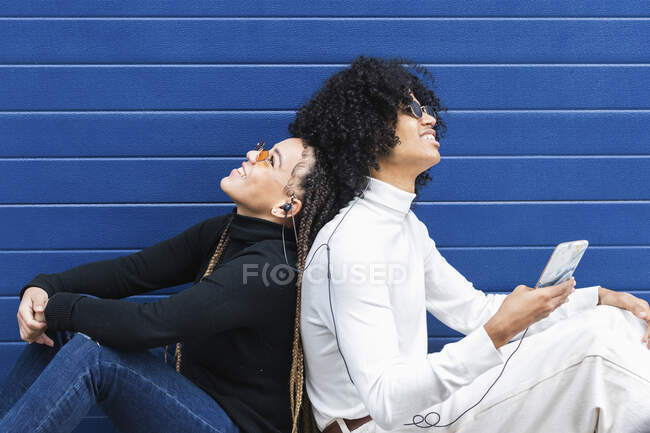 Couple listening music through in-ear headphones while sitting back to back against blue wall — Stock Photo