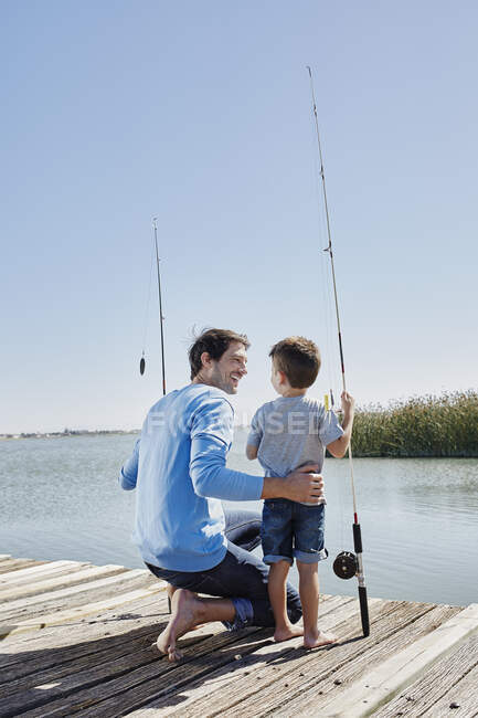 Sorridente padre guardando il figlio in piedi con la canna da pesca sul molo — Foto stock