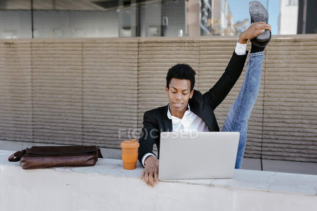 Flexibler männlicher Unternehmer sitzt mit Laptop an Stützmauer — Stockfoto