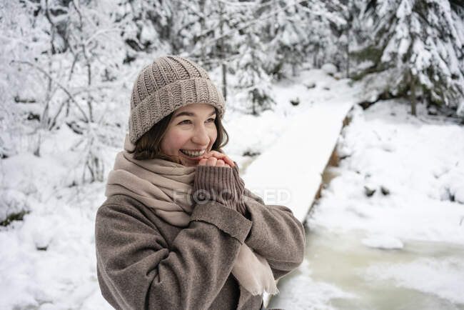 Happy woman wearing knit hat and jacket looking away while standing in forest — Stock Photo