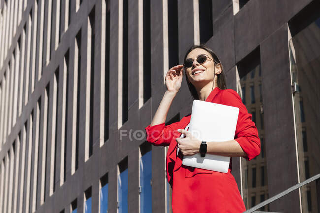 Donna d'affari sorridente con occhiali da sole che tiene il computer portatile mentre in piedi contro la costruzione — Foto stock