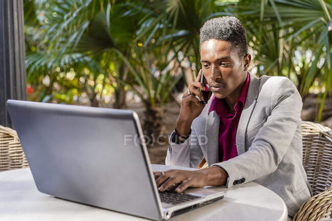 Empresario que trabaja en el ordenador portátil mientras habla a través del teléfono móvil en la cafetería - foto de stock