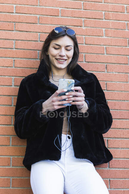 Femme souriante avec téléphone portable écoutant de la musique tout en s'appuyant sur un mur de briques — Photo de stock